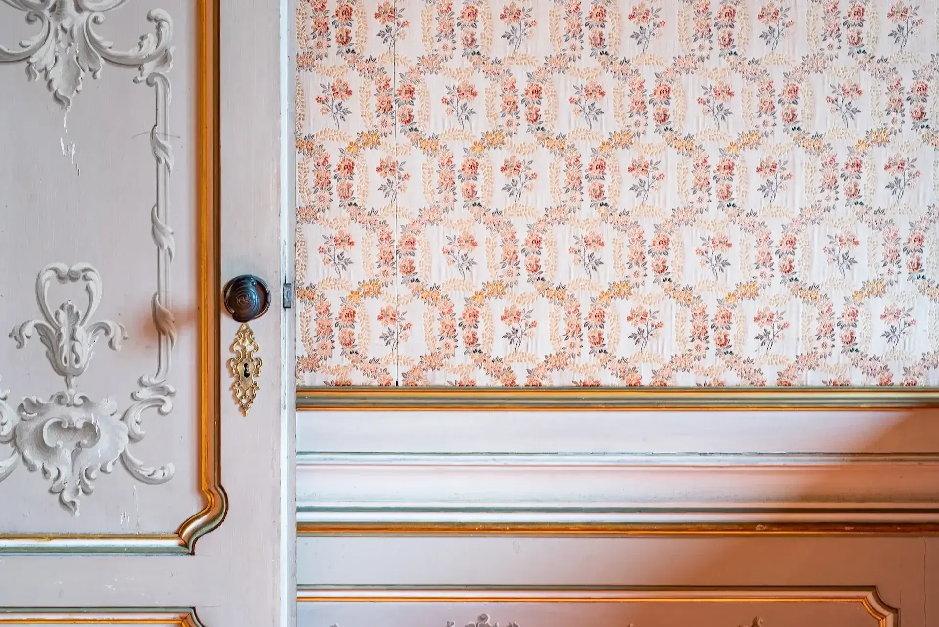 Oude deur met ornamenten en antieke deurknop, omringd door bloemetjesbehang en lambrisering met sierlijst, gerenoveerd door Walraven Bouw in Huissen voor het herstel van monumentale panden